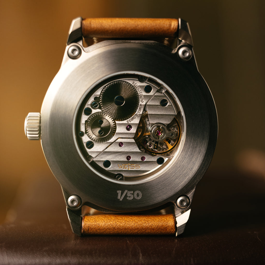Close-up of the back of a wristwatch with a brushed metal case, visible mechanical movement through the open back, and a brown leather strap on a warm brown background.
