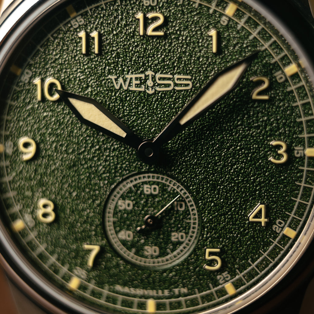 Close-up of a Weiss watch dial with a textured dark green dial, warm numerals and hands, and a small subdial at the 6 o'clock position.