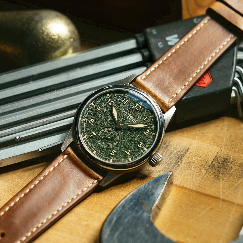 A wristwatch with a dark green dial and a brown leather strap, resting on a wooden surface with tools in the background.