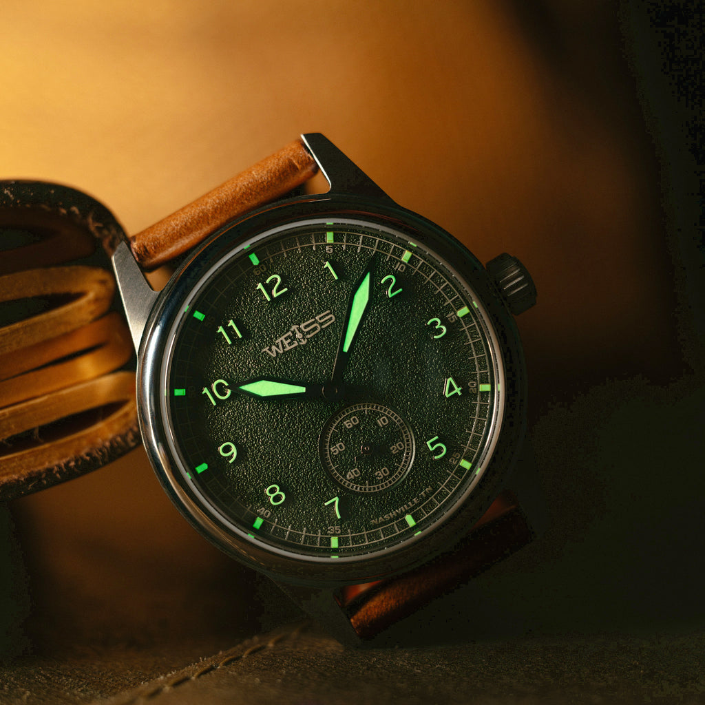 A close-up of a wristwatch with a dark green dial, bright green luminous hour markers and hands, a silver case, and a brown leather strap, resting on a leather surface with a warm brown background.