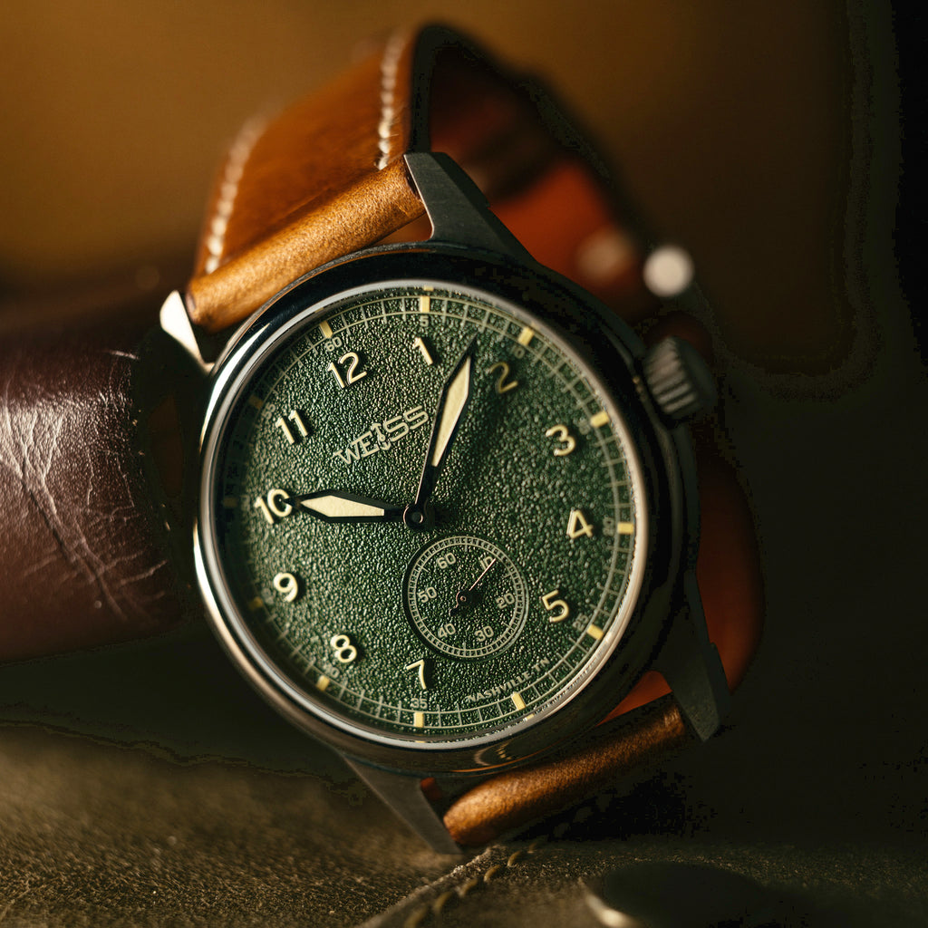 A close-up of a wristwatch with a dark green textured dial, silver case, and brown leather strap resting on a leather surface.