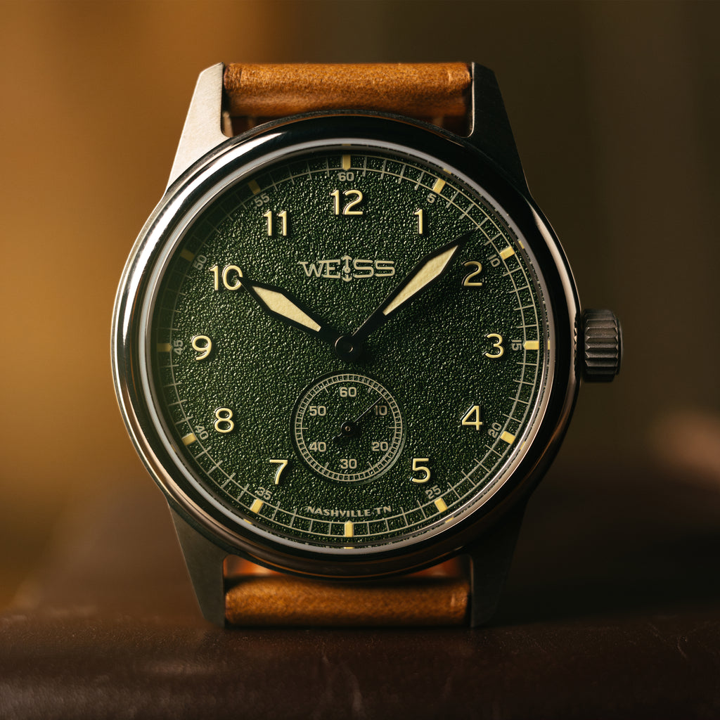 A close-up of a vintage-style wristwatch with a dark green textured dial, silver case, and a brown leather strap, resting on a dark brown surface with a warm, blurred background.