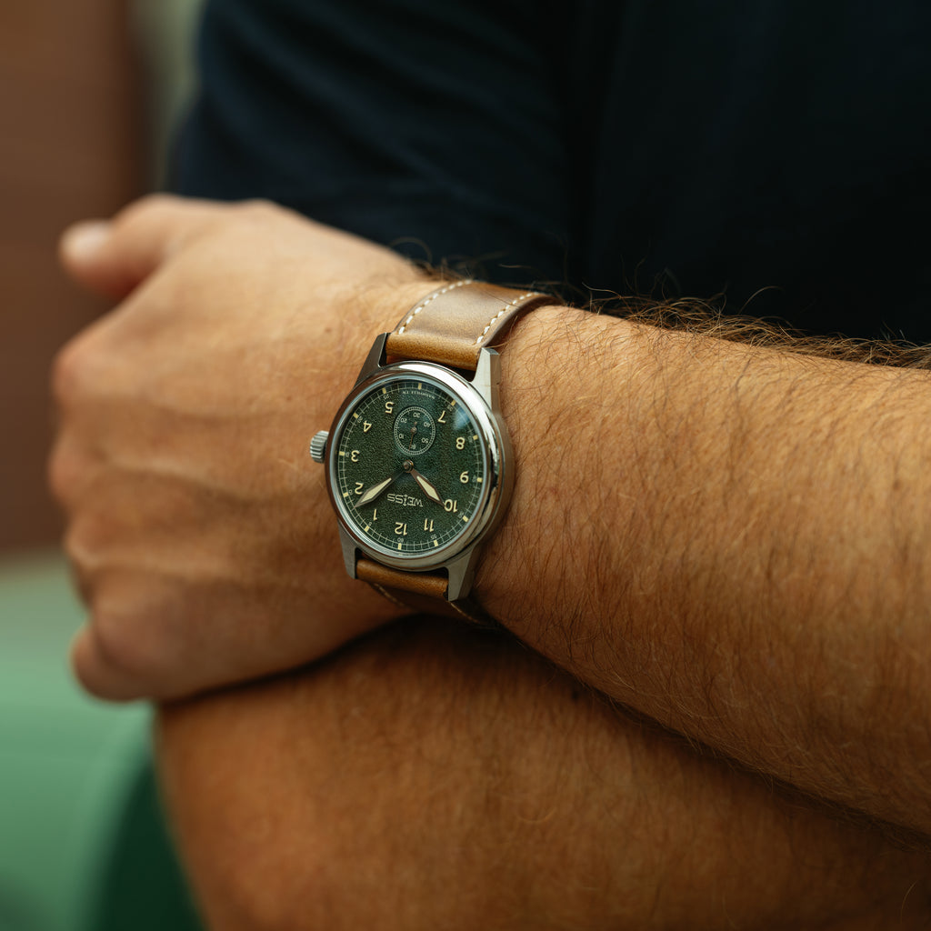 A close-up of a man's wrist wearing a vintage-style watch with a dark green dial, silver-toned case, and a light brown leather strap.