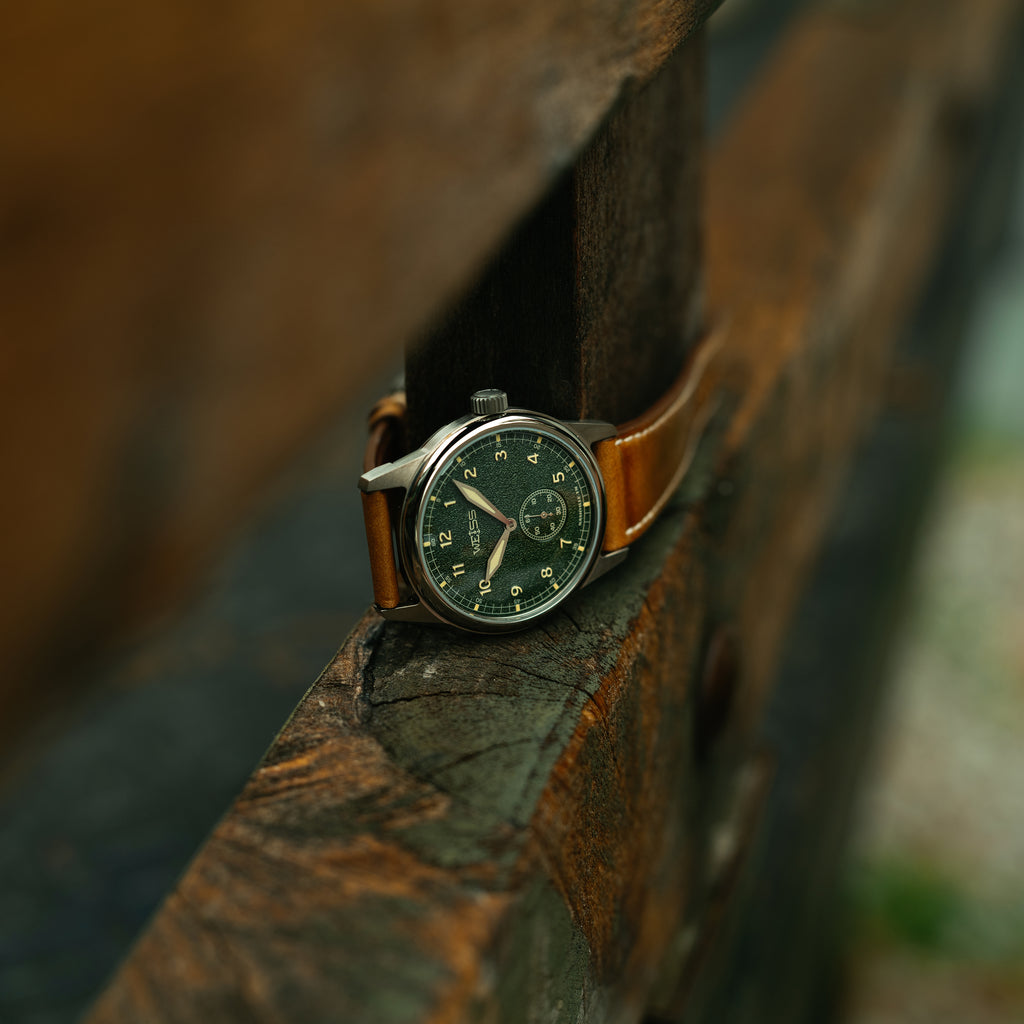 A vintage-style wristwatch with a dark green dial and brown leather strap resting on a weathered wooden surface in an outdoor setting.